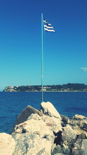 Greek flag on rocks by sea against blue sky