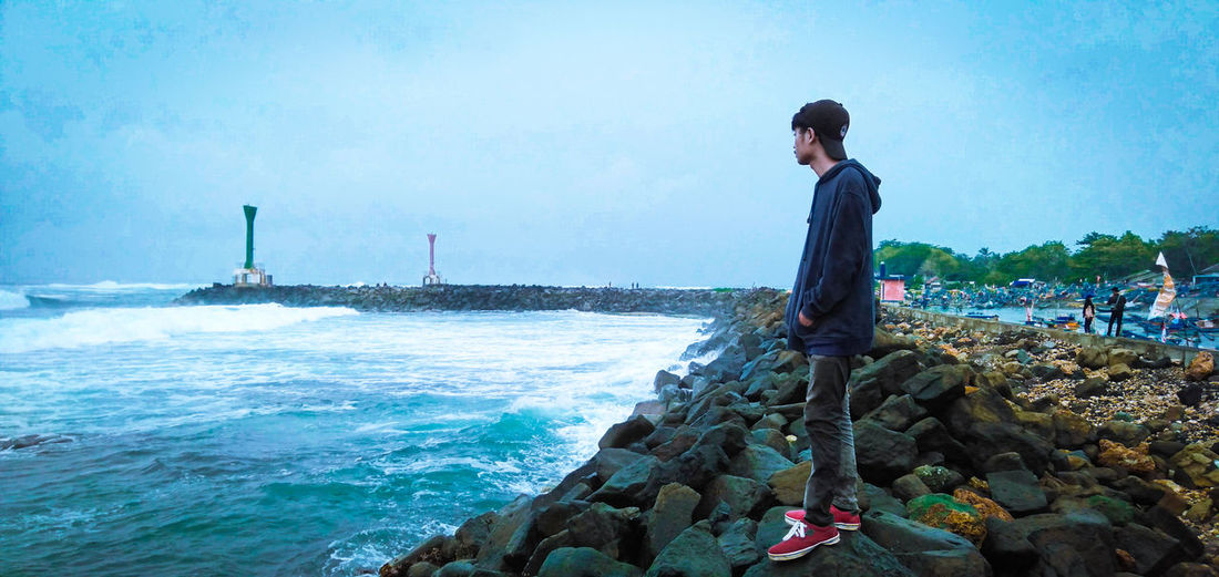 Young man standing on rock by sea against sky