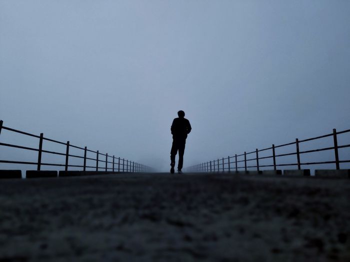 Rear view of silhouette man walking on bridge against clear sky