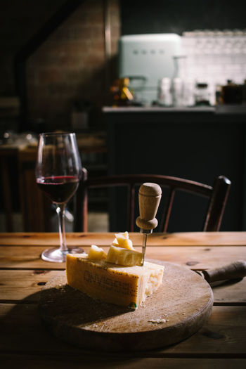 Piece of delicious cheese with knife served on wooden board placed near glass of red wine on wooden plank table in rustic bar