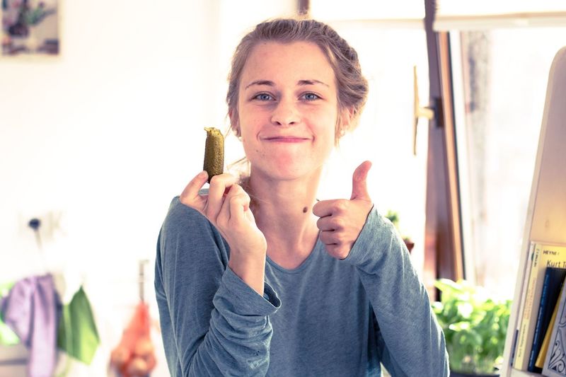 Portrait of young woman showing thumbs up while holding pickled cucumber at home
