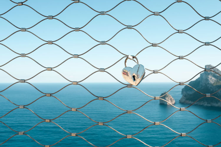 Heart shape love lock hanging on fence against mediterranean sea
