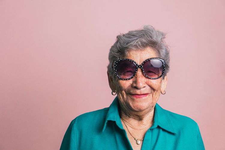 Happy modern aged female with gray hair and in trendy sunglasses on pink background in studio and looking at camera