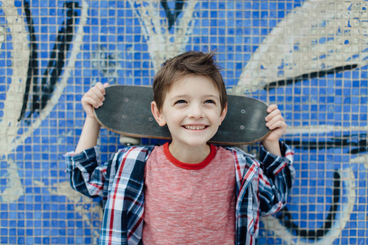 Close-up of happy boy holding skateboard while standing by wall