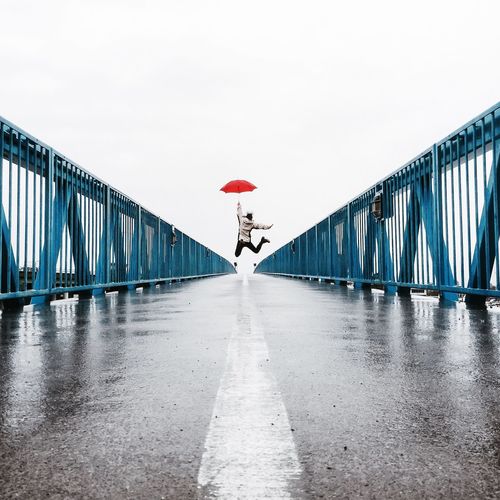 Man with umbrella jumping on bridge against clear sky