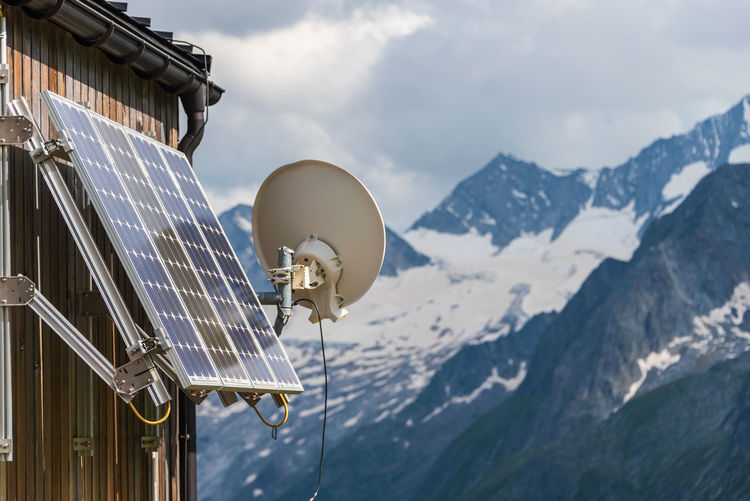Pv solar panels and satellite dish antenna at the wall of a wooden building with snowy mountains