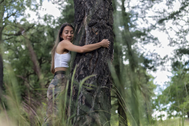 Tranquil female in casual wear with closed eyes hugging thick tree trunk in forest with blurred green grass during hiking