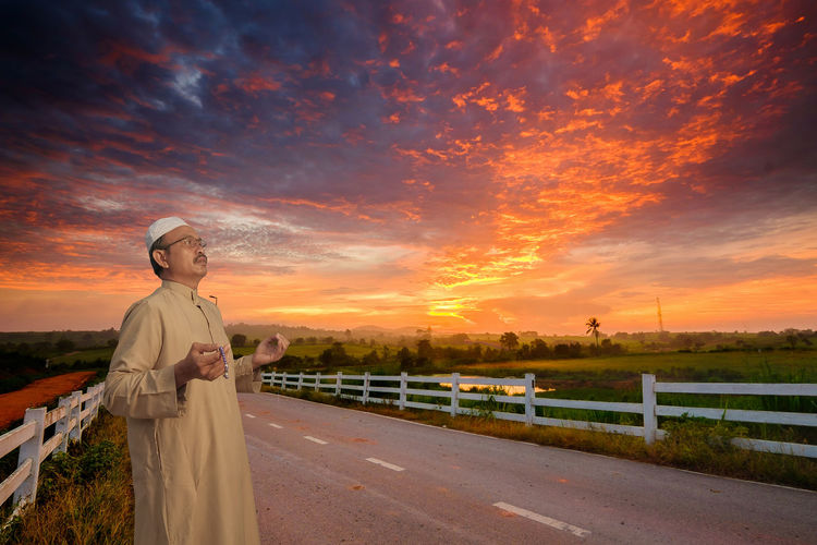 Mature man wearing taqiyah praying while standing on road against dramatic sky during sunset