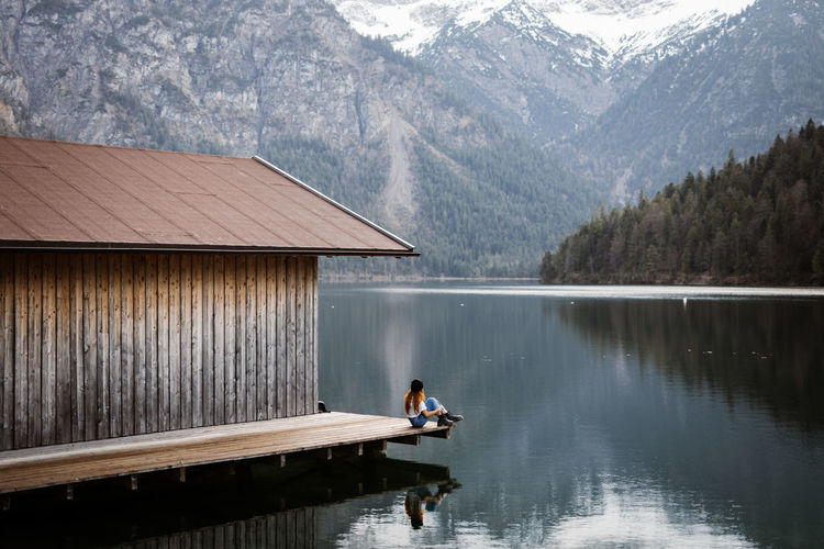 Side view of unrecognizable female sitting on wooden quay and enjoying scenery of calm pond and mountains covered with snow in winter