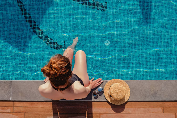 Young woman sitting on the edge of the swimming pool with her straw hat lying near, top view
