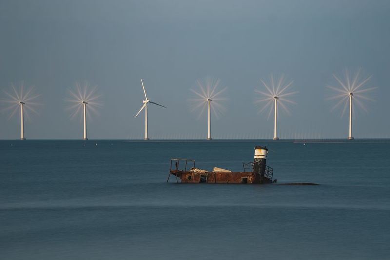Abandoned boat by wind turbines on sea shore against sky