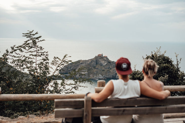 Rear view of couple sitting on bench by railing against sky
