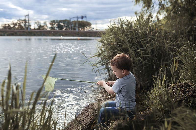 Blond-haired boy in blue clothes sits on lake and catches green mud in net, little fisherman