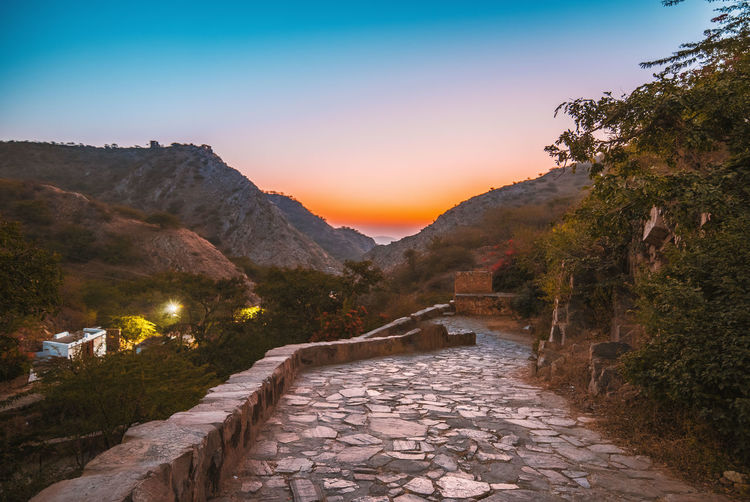 Footpath leading towards mountains against sky during sunset