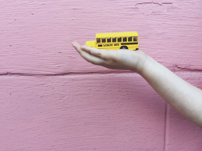 Cropped hand holding toy school bus against pink wall