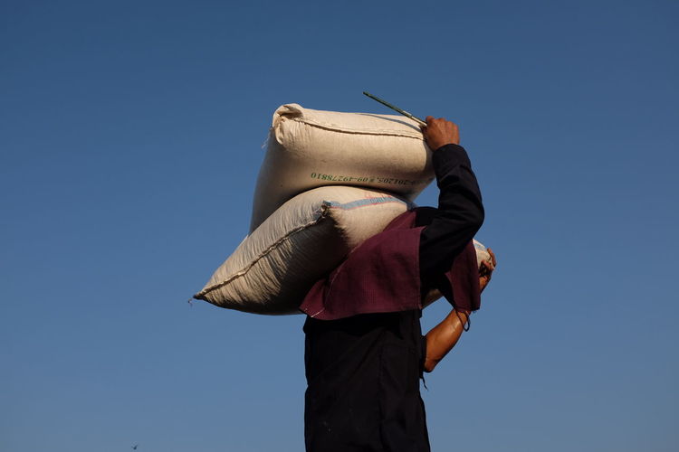 Side view of man carrying sack while standing against blue sky