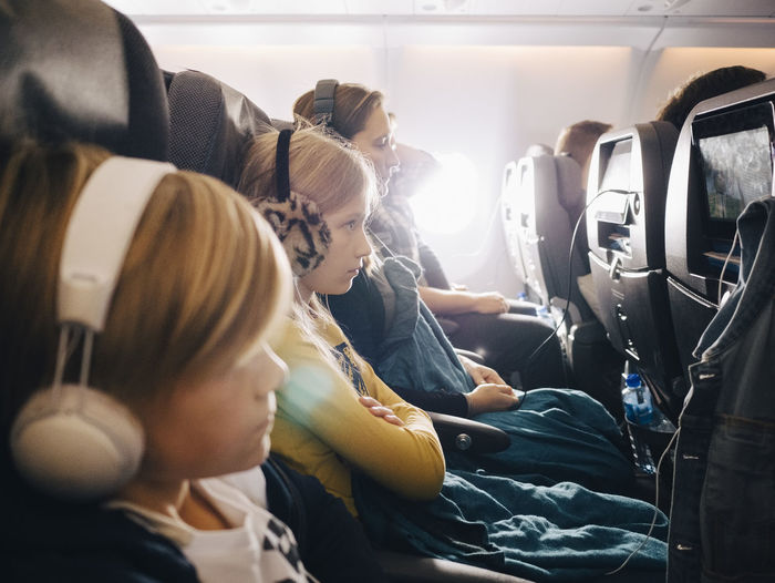 Side view of mother and children sitting in airplane while using headphones