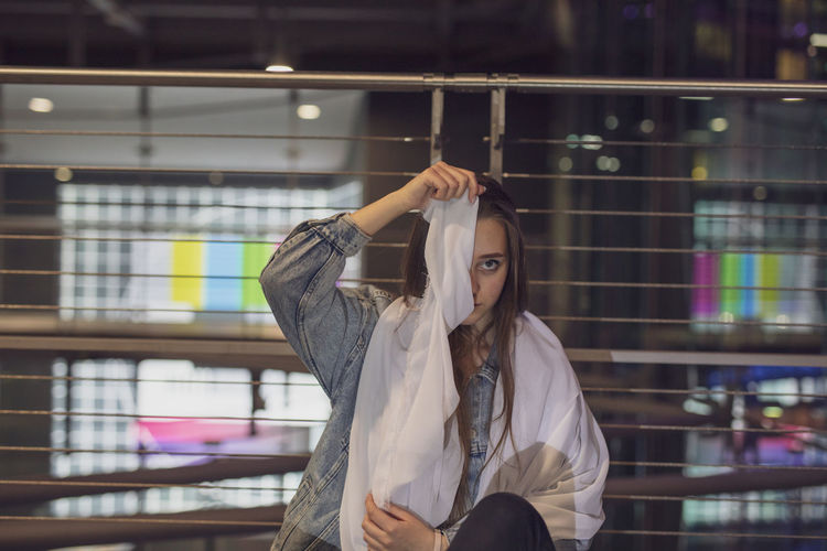 Portrait of young woman holding fabric over face against railing