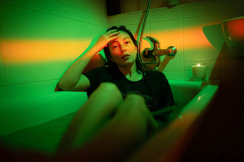 Young depressed asian woman in t-shirt sitting in bathtub under neon light. mental health problems