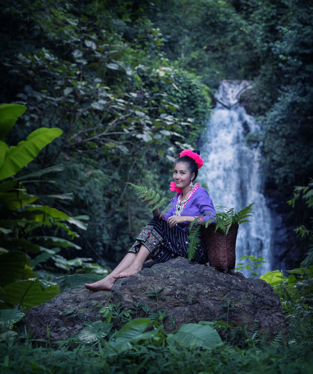 Full length portrait of young woman sitting on rock against waterfall