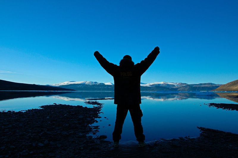 Full length rear view of silhouette man with arms raised standing on lakeshore against clear blue sky