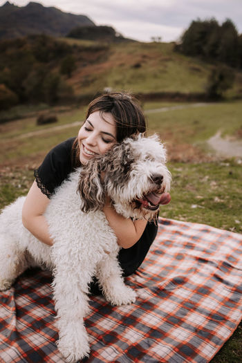Candid female owner cuddling fluffy spanish water dog while sitting on blanket in mountains and enjoying weekend together