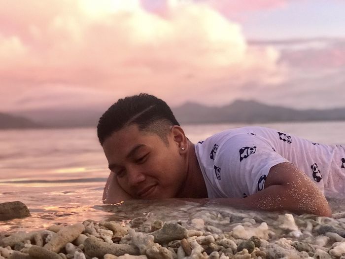 Portrait of young man relaxing on beach against sky during sunset