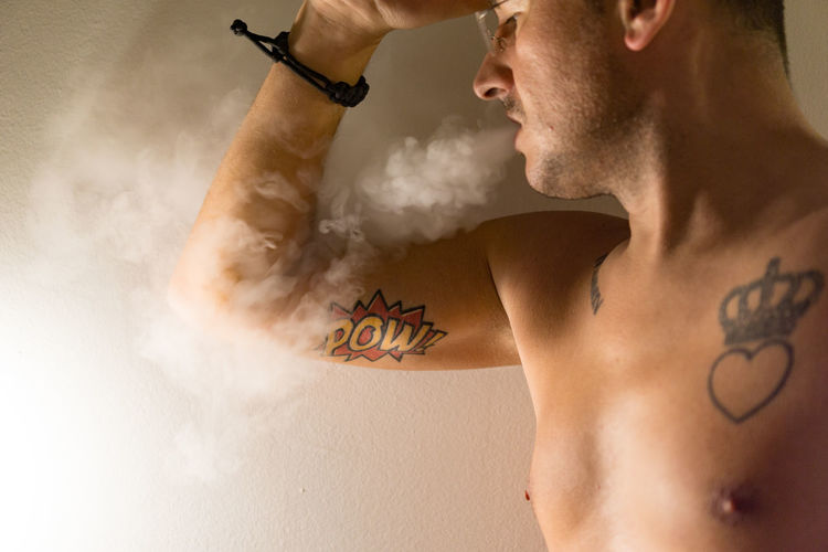 Midsection of shirtless man exhaling smoke against wall