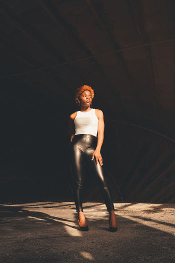 Full body of confident african american female in trendy outfit and high heels standing in dark tunnel with bright sunlight