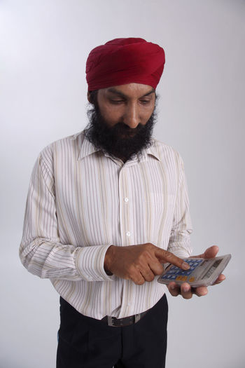 Bearded man wearing red turban using calculator against white background