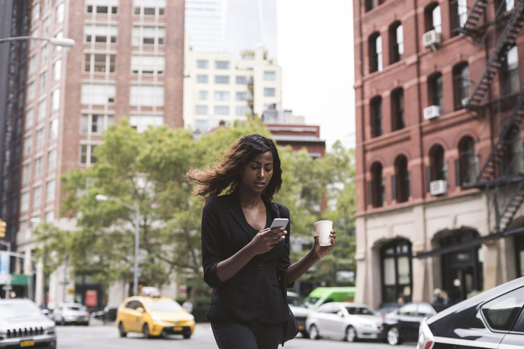 Businesswoman with coffee using smart phone while walking on city street