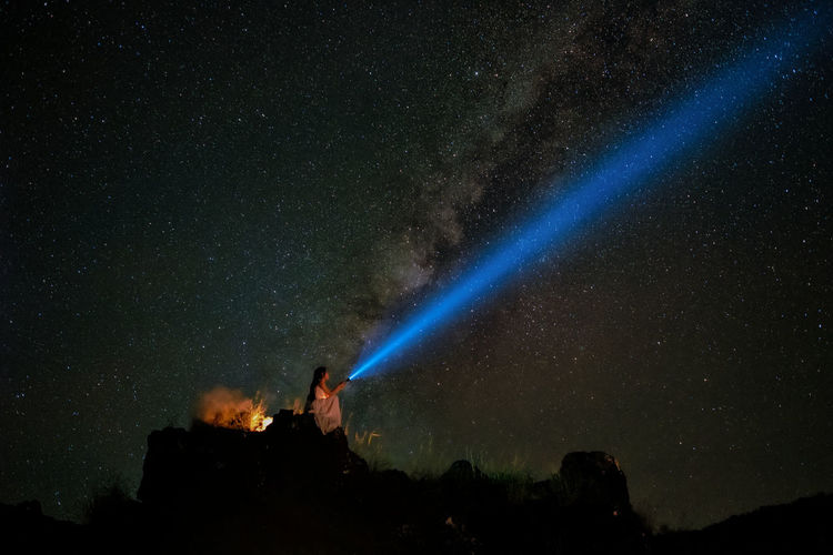 Low angle view of woman with flashlight sitting on cliff against sky at night