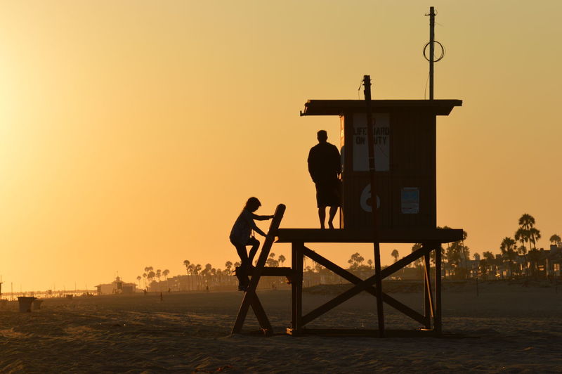 Silhouette lifeguards on hut at beach against orange sky
