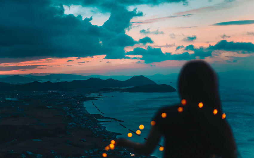 Rear view of silhouette woman with illuminated string light standing by sea against sky