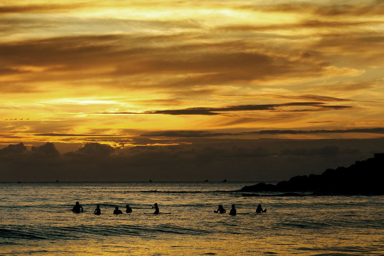 Silhouette people enjoying in sea against cloudy orange sky during sunset