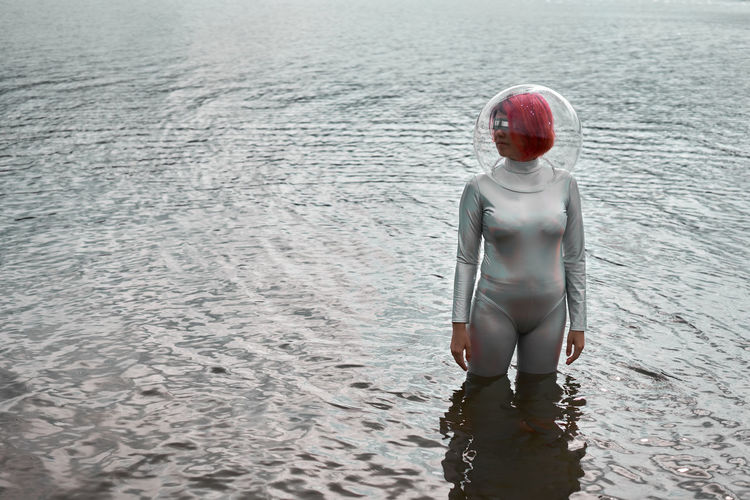 Futuristic young red haired female in silver space suit and glass helmet walking out of flooding river
