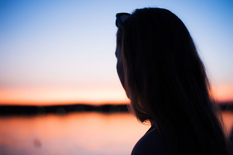 Side view of silhouette teenage girl standing by lake against sky during sunset