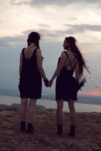 Two woman in black dresses stay on the mountain with a bouquet of flowers in summer at sunset
