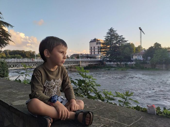 Thoughtful boy sitting on retaining wall by river in city