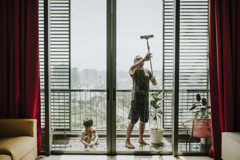 Father with son cleaning windows in balcony seen through glass