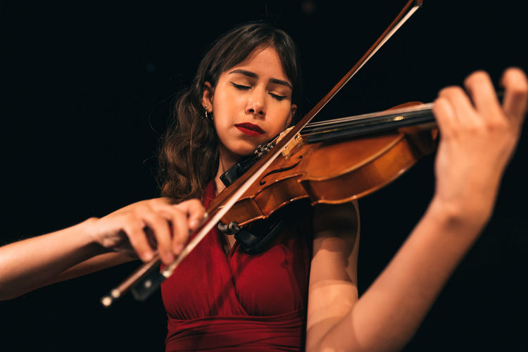 Woman in red dress playing violin with closed eyes during concert on stage in dark theater
