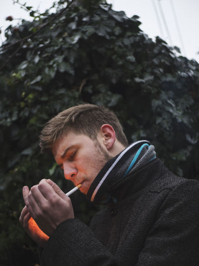 Low angle view of young man smoking while standing against trees in winter