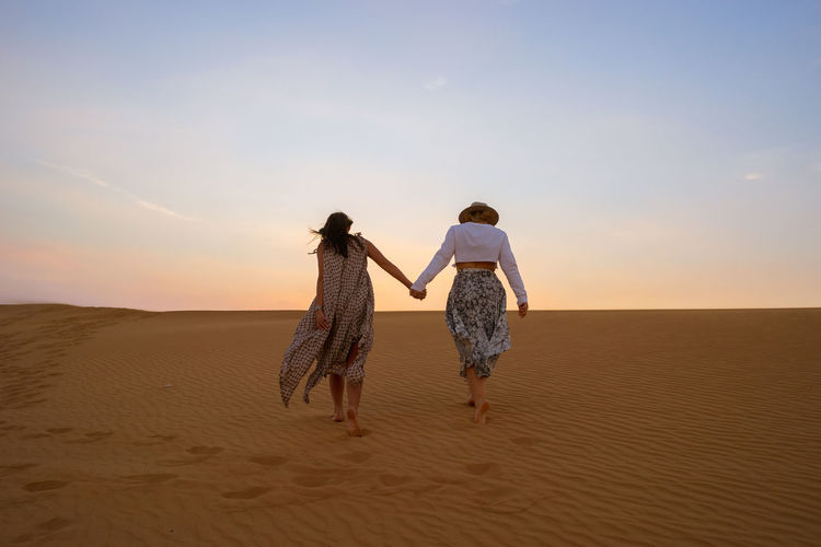 Full length rear view of women holding hands while walking on beach