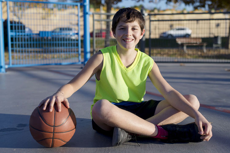 Portrait of smiling boy holding ball while sitting on basketball court