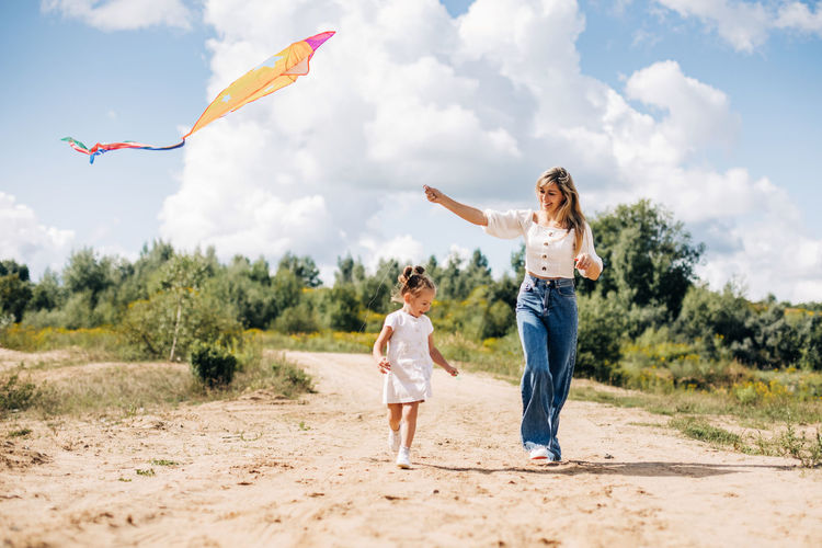 A laughing little girl and her mother run along the road to the field and fly a kite.
