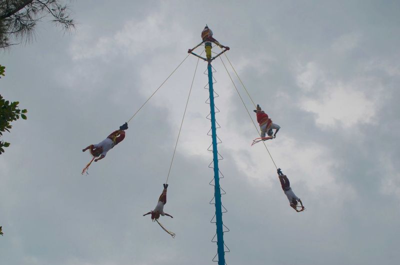 Low angle view of men performing volador against cloudy sky