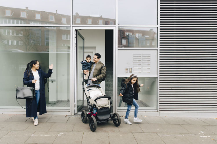 Mother waving to father and children with stroller while walking on sidewalk in city
