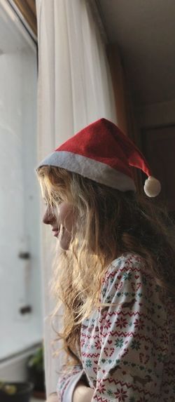 Profile portrait of teenage girl wearing red santa's hat and looking at the window.