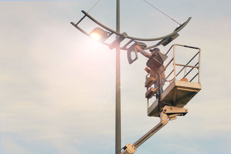 Low angle view of man repairing street light while standing on cherry picker against sky