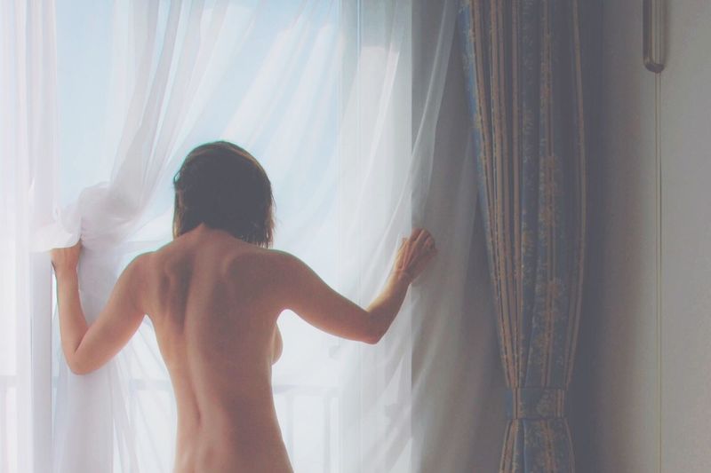 Rear view of naked woman standing by curtains at home